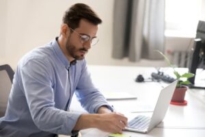 Mann im Hemd sitzt am Schreibtisch mit Laptop und Stift