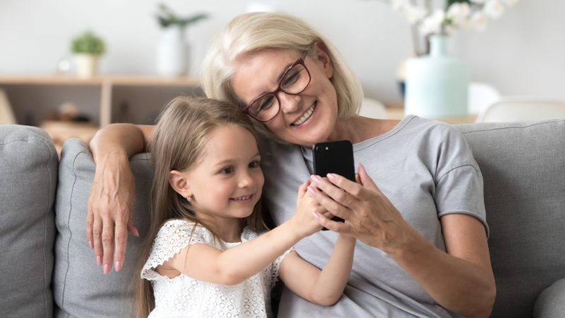 Enkelin erklärt auf dem Sofa sitzend ihrer Oma, wie das Handy funktioniert. Sie sind beide glücklich.