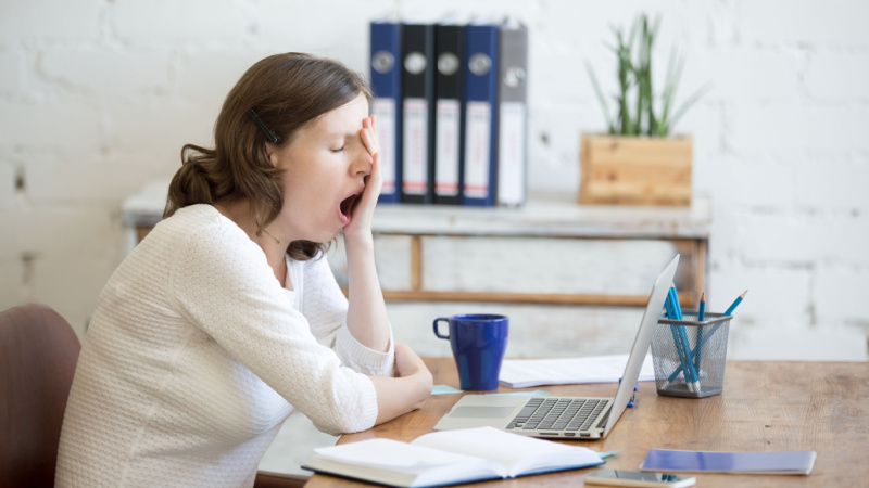 Ausreden, warum man nicht in die Arbeit kommt: Frau sitzt am Schreibtisch mit Kaffee und Laptop und gähnt.