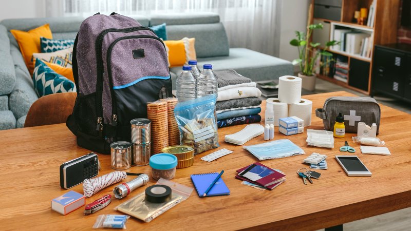 Die Ausrüstung eines Notfallrucksacks ist auf einem Tisch ausgebreitet. Dort liegen beispielsweise Wasserflaschen, Ausweisdokumente, Erste-Hilfe-Set, Handy, Taschenlampe und vieles mehr.