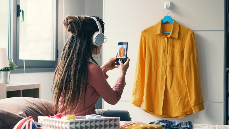 Frau sitzt auf ihrem Bett und fotografiert mit ihrem Handy eine gelbe Bluse ab.