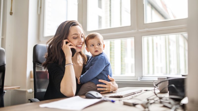 Junge Frau, die mit ihrem Sohn am Tisch sitzt und im Büro mit dem Handy telefoniert.