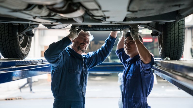 Kfz-Mechaniker und ein Lehrling arbeiten unter einem Auto